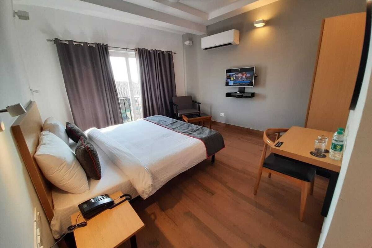 A hotel room with a bed, bedside table, chair, desk, TV, and an air conditioning unit.
