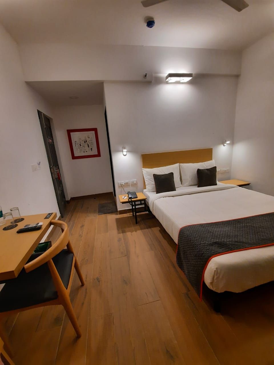 Habitación de hotel con cama, mesita de noche, escritorio con silla, una pequeña mesa con silla, y una lámpara fijada a la pared.