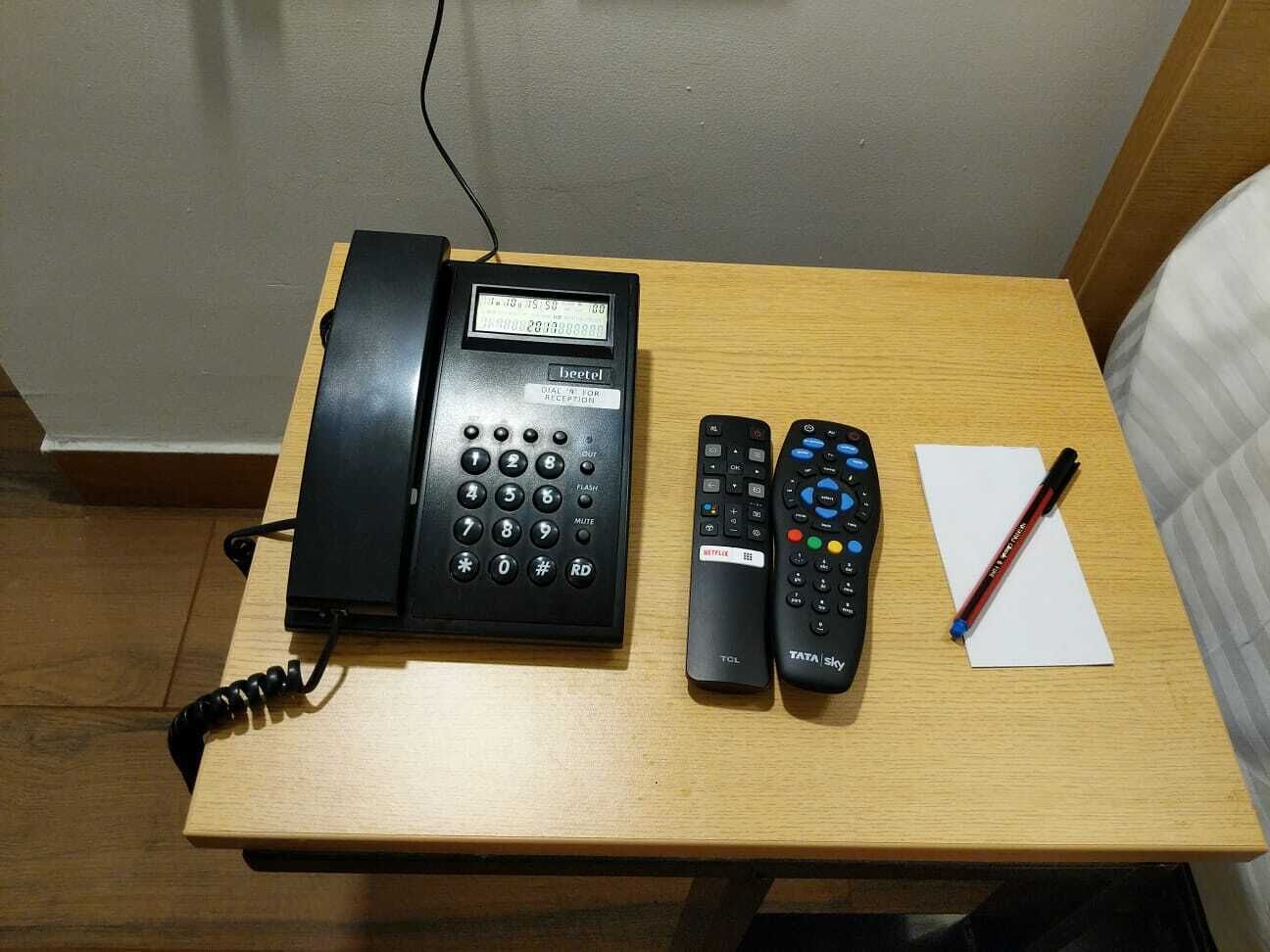 Escritorio de madera con un teléfono negro, dos controles remotos y un bolígrafo sobre un papel.