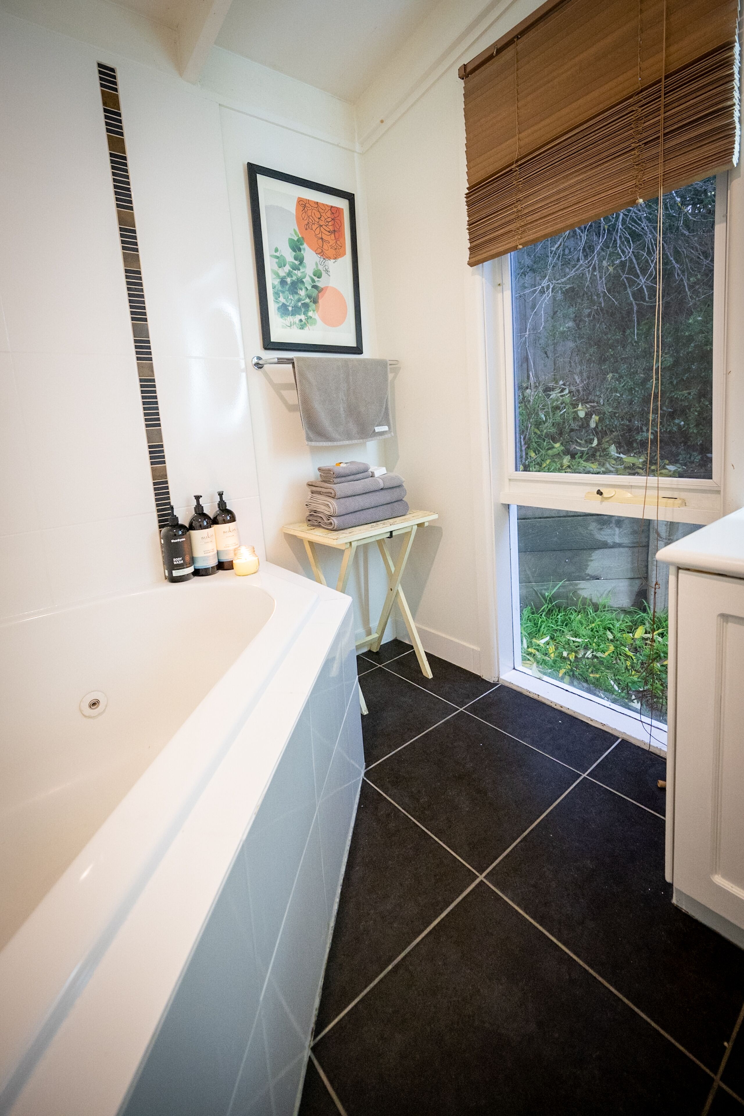A modern bathroom with a white bathtub, a small table with towels, and a window with a view of greenery.