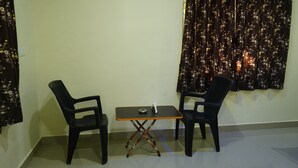 Une petite pièce avec deux chaises noires et une table, une fenêtre ornée de rideaux à motifs floraux et un mur uni.