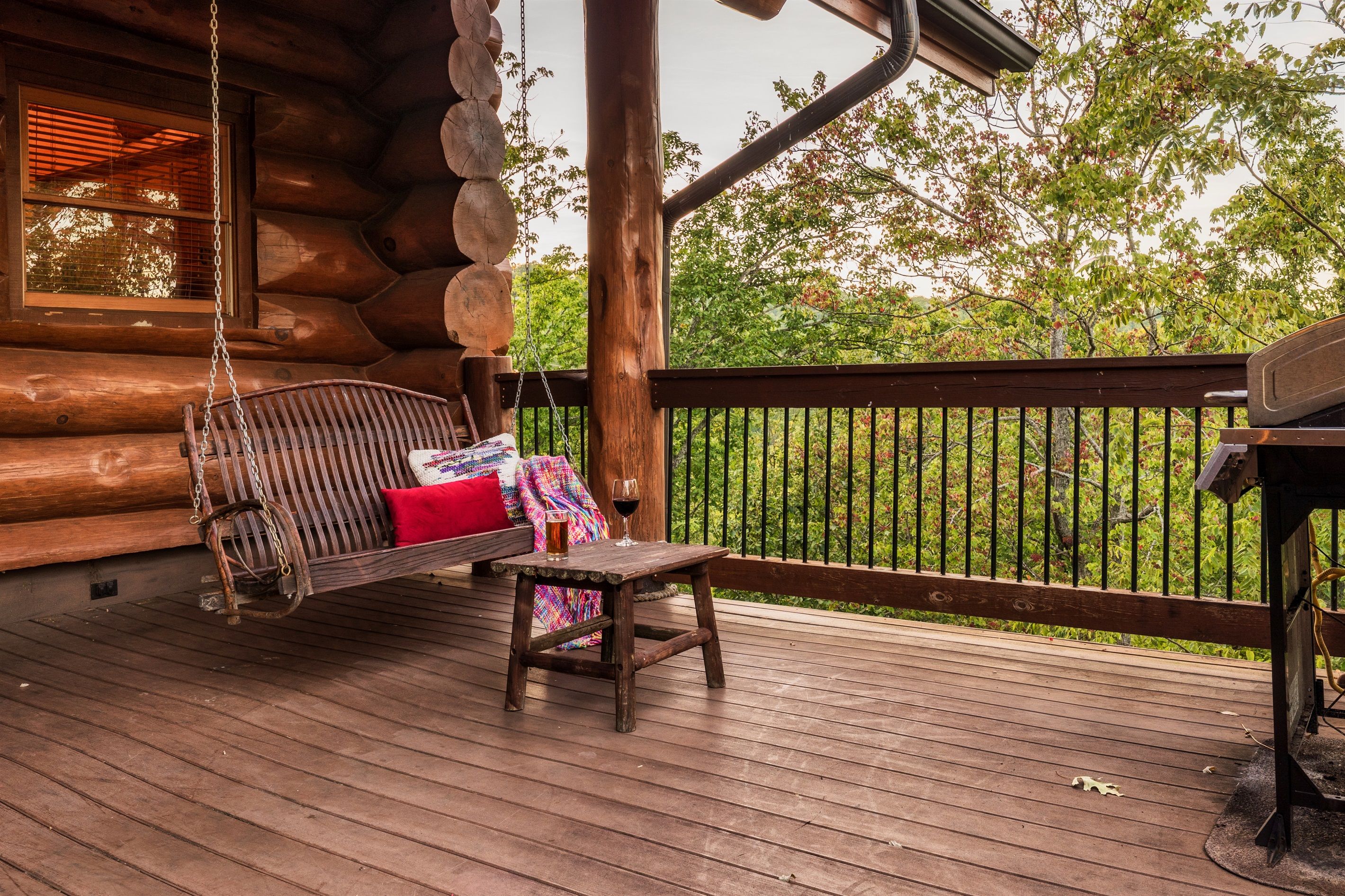Une cabane en rondins avec une terrasse en bois, une balançoire suspendue, une petite table avec un verre de vin et une vue sur la verdure.