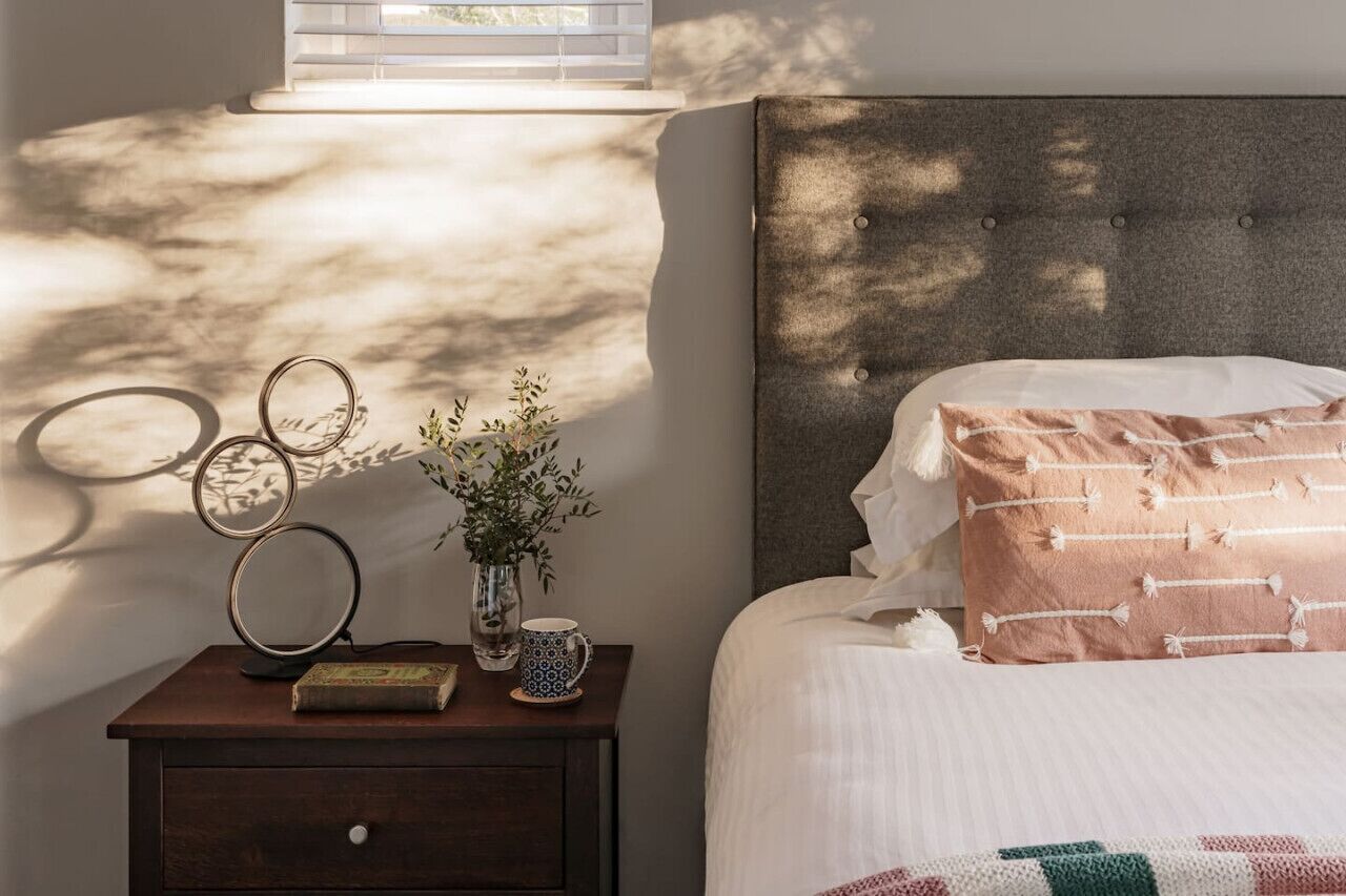 A neatly made bed with a patterned pillow, a bedside table with a book, a vase with plants, and wall art.