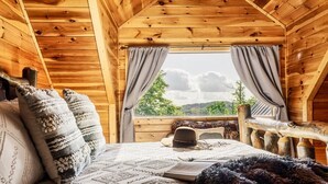 A cozy wooden cabin room with a bed, pillows, a hat, and a window with curtains.