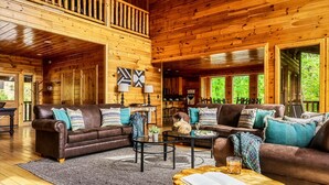 A spacious wooden cabin with a high ceiling, furnished with leather sofas and a coffee table, featuring a view of the surrounding forest.