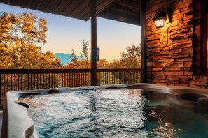 A hot tub with a wooden deck and a stone wall, overlooking a forested area with autumn foliage.