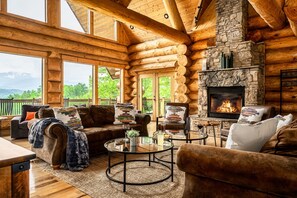 Interior de una cabaña de troncos con chimenea de piedra, muebles de madera y ventanas amplias que ofrecen vistas al exterior.