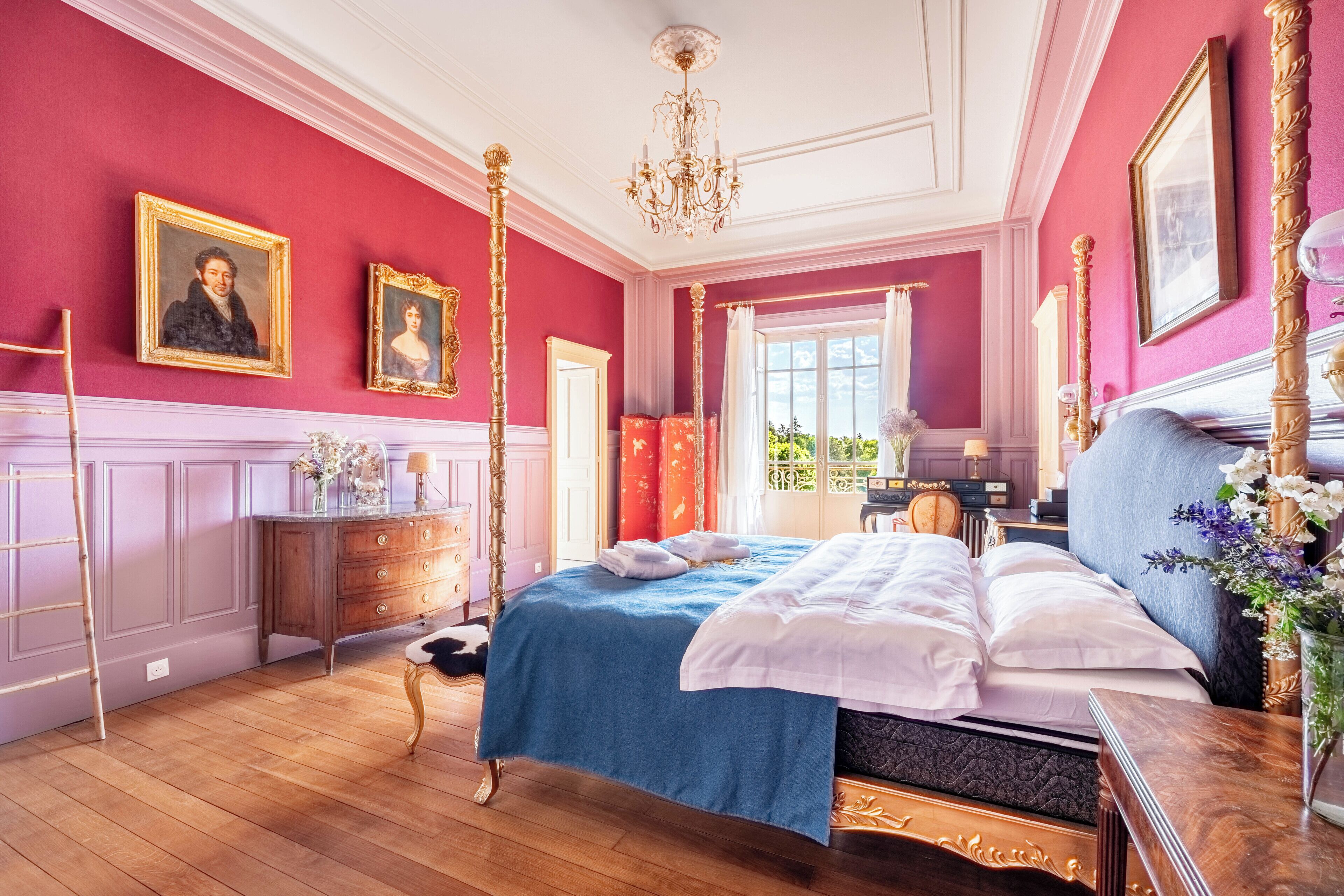 A room with a four-poster bed, a wooden dresser, a chandelier, and two framed portraits on the wall.