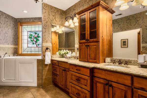 Un baño con una bañera independiente, muebles de madera, un espejo grande y un ventanal decorativo con diseño floral.