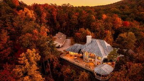 Una casa grande de varios pisos rodeada de un bosque frondoso, con una chimenea destacada y un balcón con vistas al paisaje.