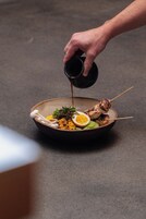A hand pouring a liquid from a small black cup onto a bowl containing food.