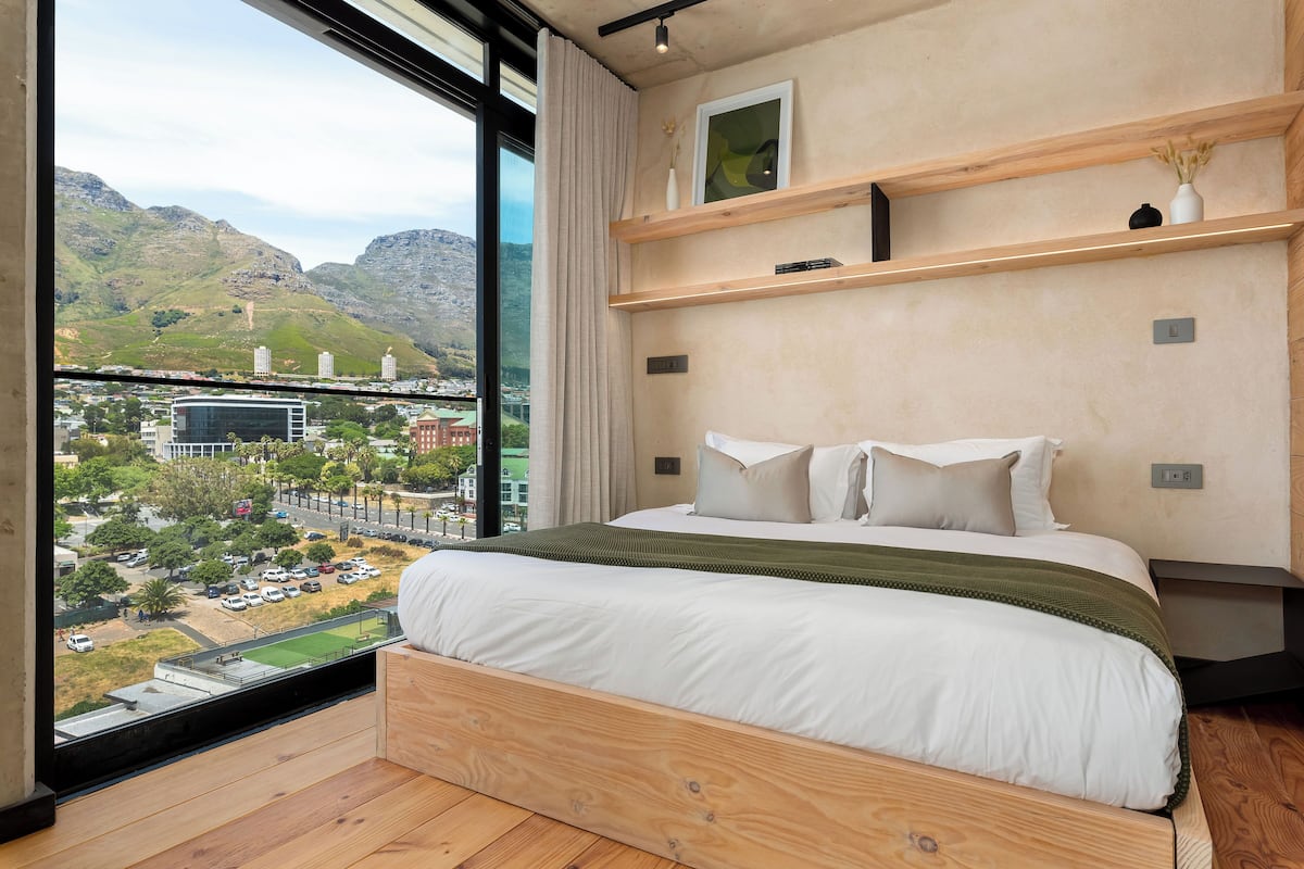 Un dormitorio con un ventanal grande, estructura de cama de madera y vistas a un paisaje urbano y montañas.
