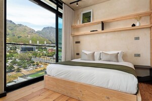 Un dormitorio con un ventanal grande, estructura de cama de madera y vistas a un paisaje urbano y montañas.