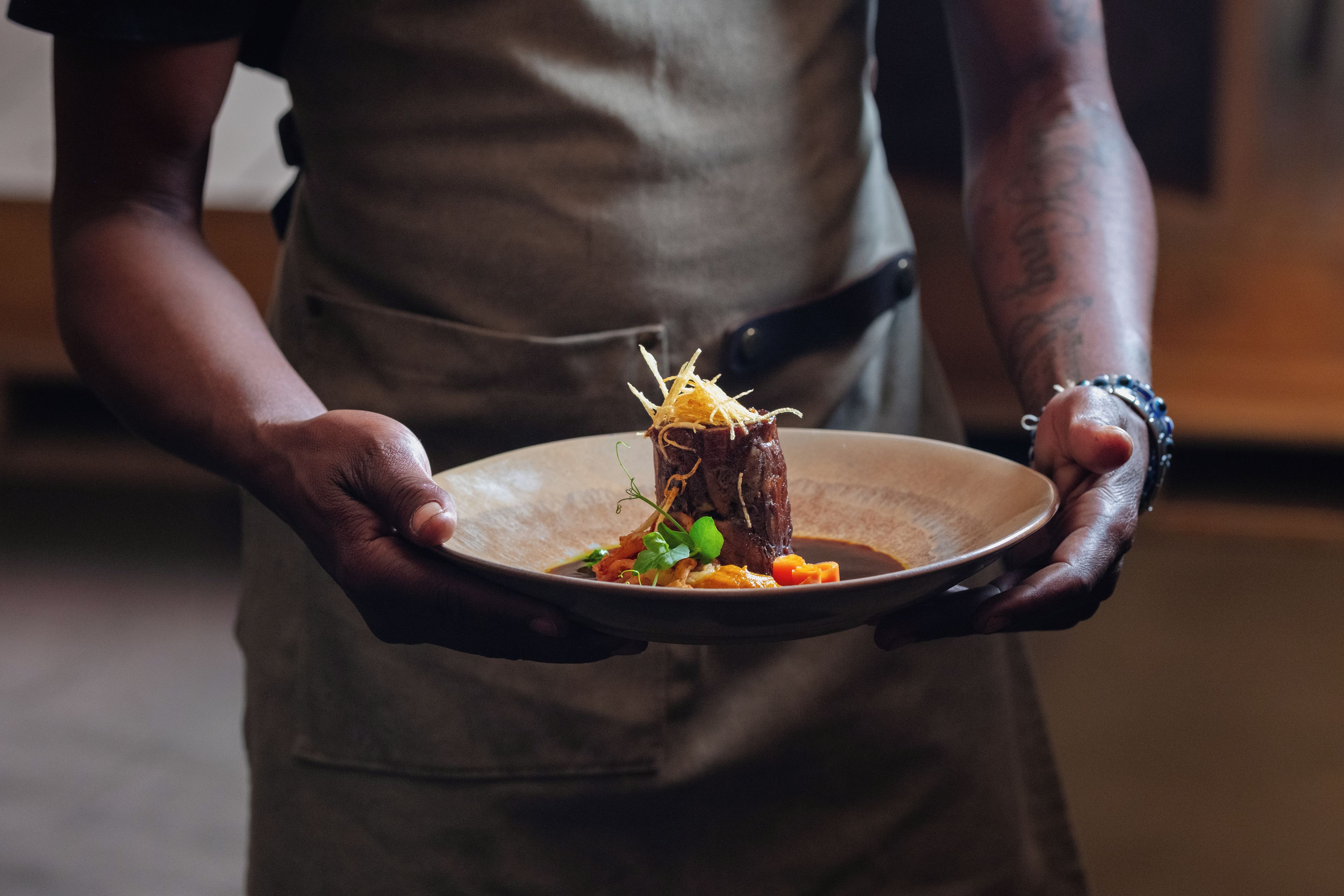 A person holding a plate with a dish that includes meat, vegetables, and a garnish.