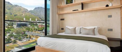 A bedroom with a large window, a bed, a wooden headboard, a bedside table, and a view of a cityscape and mountains.