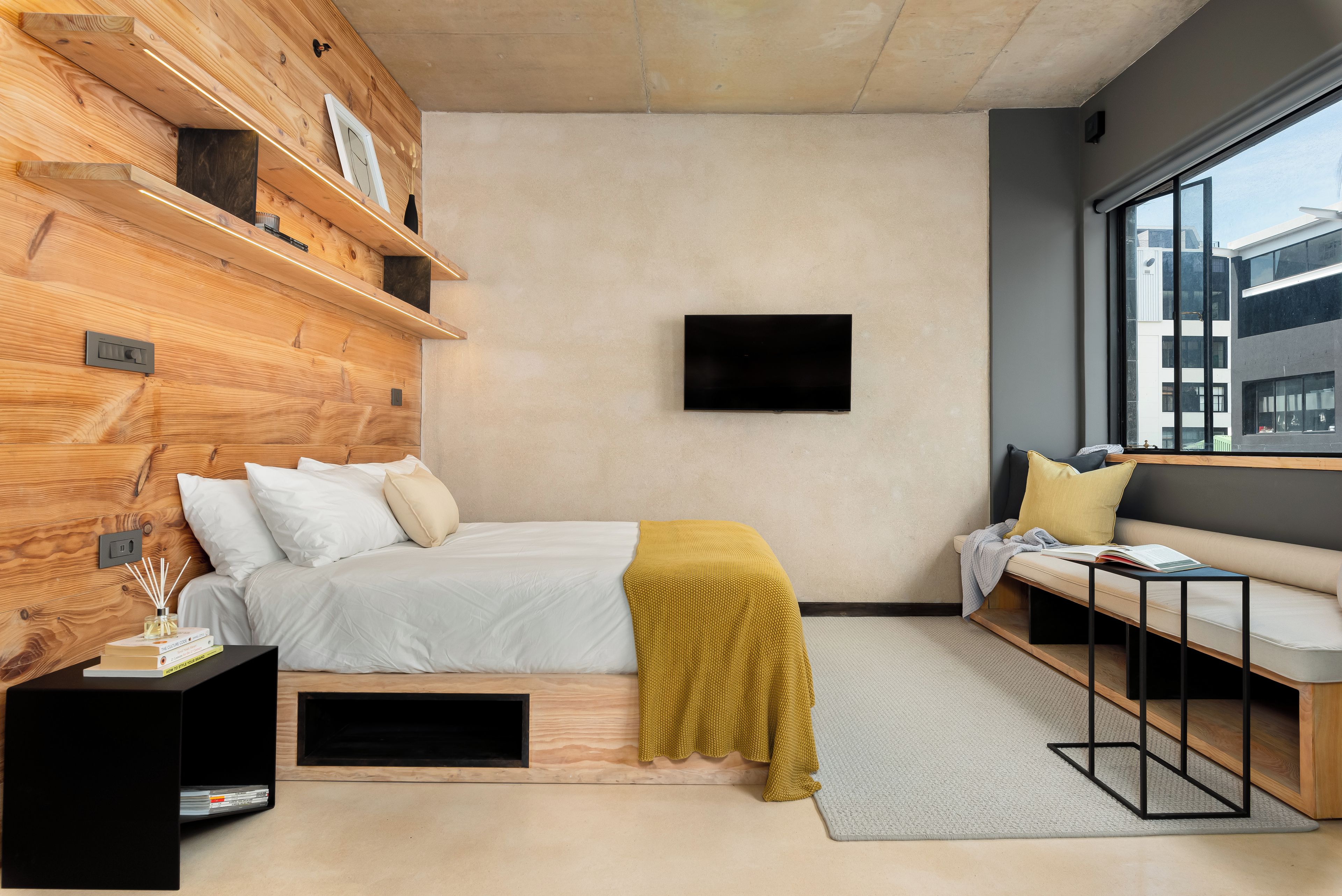 Un dormitorio moderno con cabecera de madera, cama con ropa blanca, un cojín amarillo, un televisor montado en la pared, un banco con almohadones y un ventanal grande.