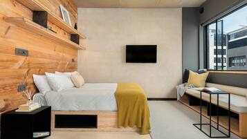 Un dormitorio moderno con cabecera de madera, cama con ropa blanca, un cojín amarillo, un televisor montado en la pared, un banco con almohadones y un ventanal grande.