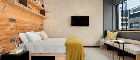A modern bedroom with a wooden headboard, a bed with white bedding, a yellow throw, a TV mounted on the wall, a bench with cushions, and a large window.