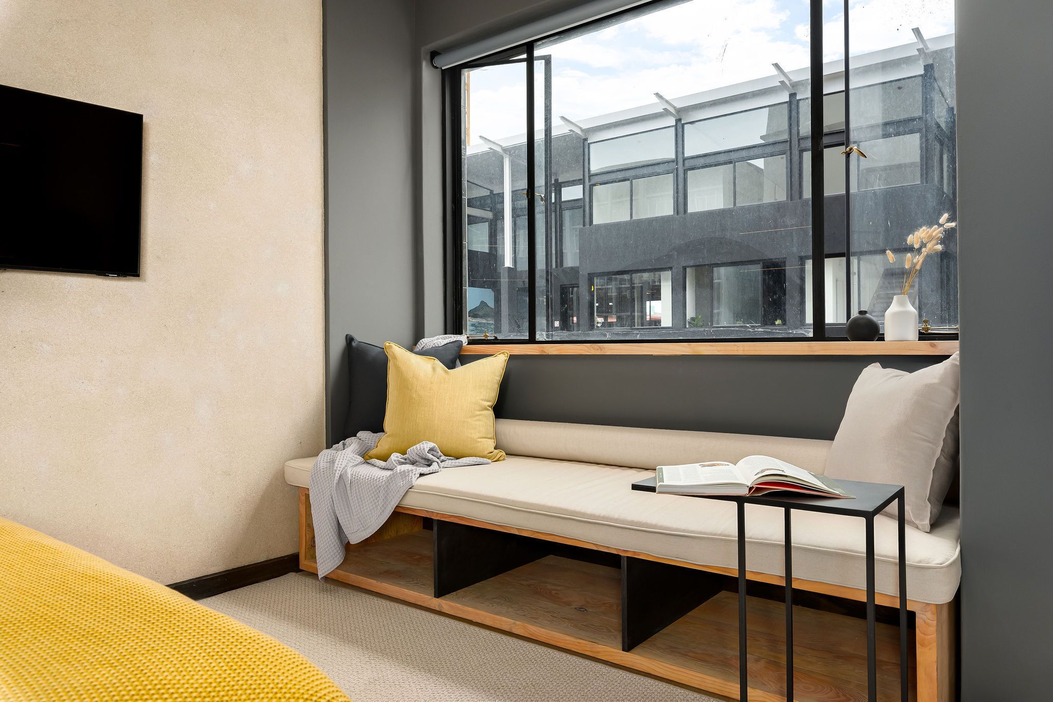 Une chambre moderne dotée d’une grande fenêtre, d’un banc avec des coussins, d’un téléviseur à écran plat et d’une petite table sur laquelle est posé un livre.