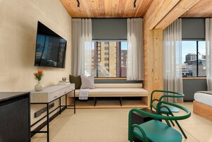 A modern hotel room with a large window, a sofa, a desk, and a TV mounted on the wall.