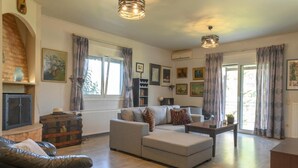 A living room with a sofa, a coffee table, and a fireplace. There are curtains, framed pictures on the wall, and a window with a view. - Crete Getaway 3Bd Apt w Garden and View (Malevizi)
