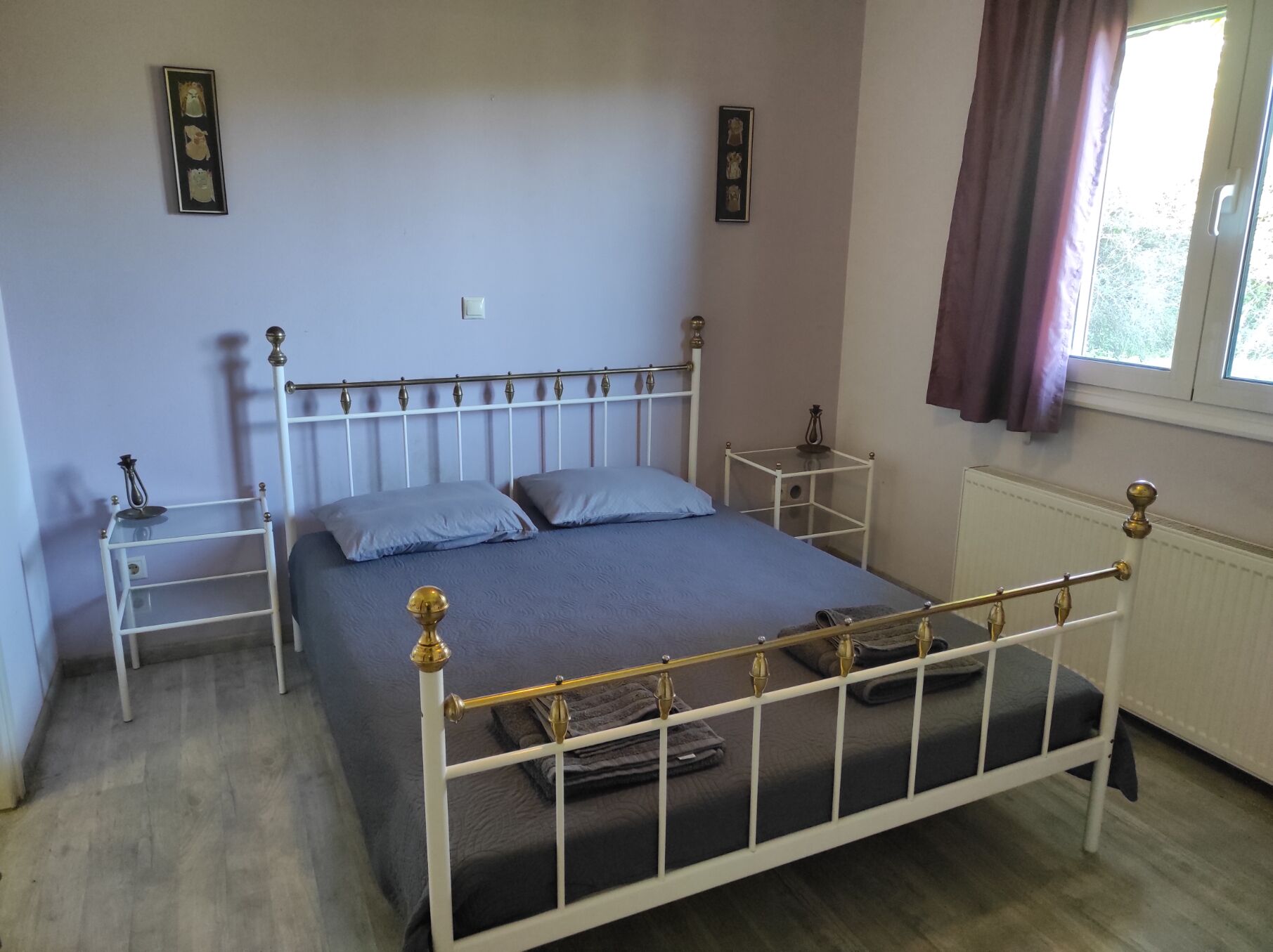 Een slaapkamer met een bed met wit metalen frame, twee nachtkastjes, een wandlamp en foto’s aan de muur, plus een raam met gordijnen.
