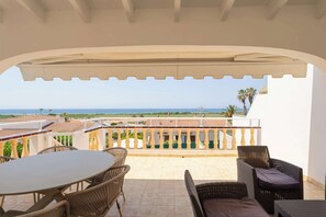 Ein überdachter Außenbereich mit Tisch und Stühlen, Blick auf das Meer und ein Balkon mit Geländer.