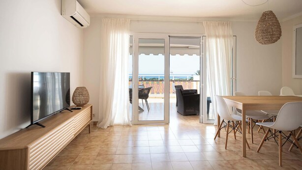A modern living room with a TV, a wooden bench, a dining table with chairs, and a view of the sea through a glass door. - Villa Ocean Breeze - Steps away from the Beach (San Jaime Mediterráneo)