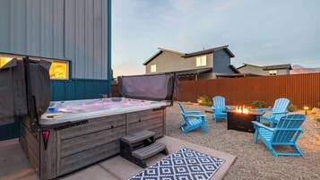 An outdoor hot tub area with a wooden bench, blue Adirondack chairs, and a fire pit.