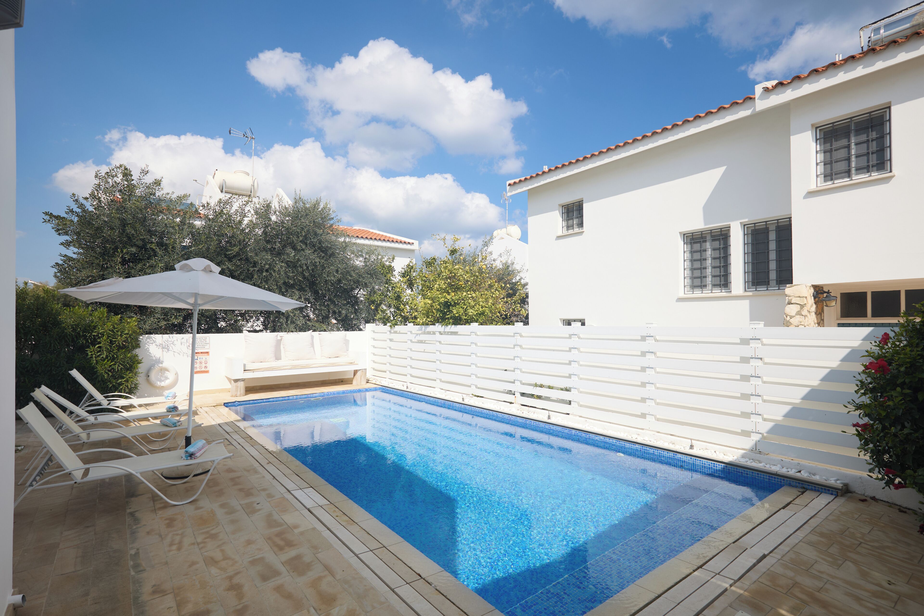 A swimming pool with lounge chairs and a white umbrella, surrounded by a white fence and a building with a tiled roof.