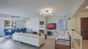 A modern living room with a white sofa, dining table with blue chairs, a TV, and a fireplace. - Protaras Serifos Villa Sv07 (Paralimni)