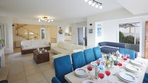 A spacious living area with a dining table set for a meal, a sofa, and a staircase leading to an upper level. - Protaras Serifos Villa Sv07 (Paralimni)