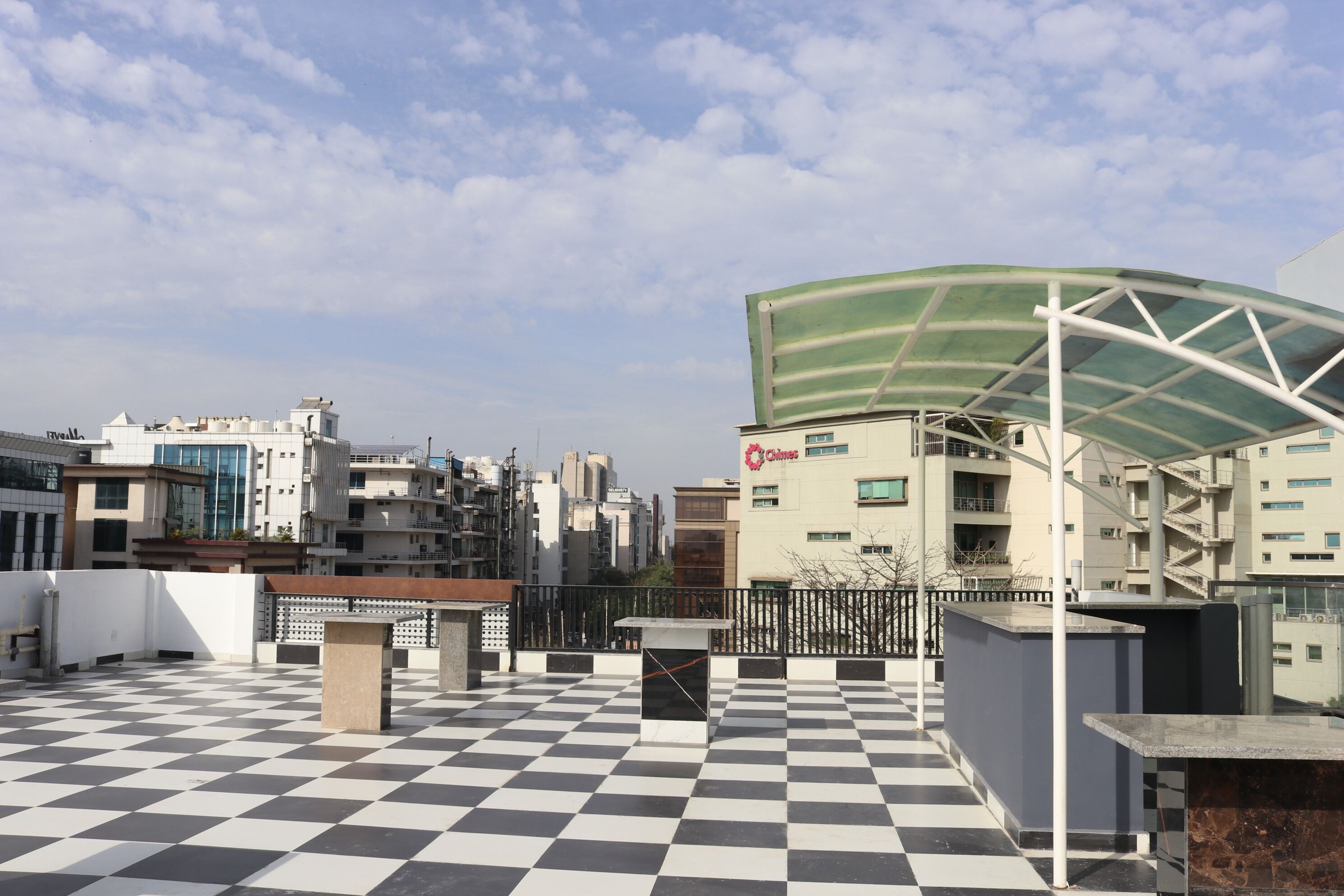 A rooftop terrace with a checkered floor, a covered seating area, and a view of surrounding buildings.