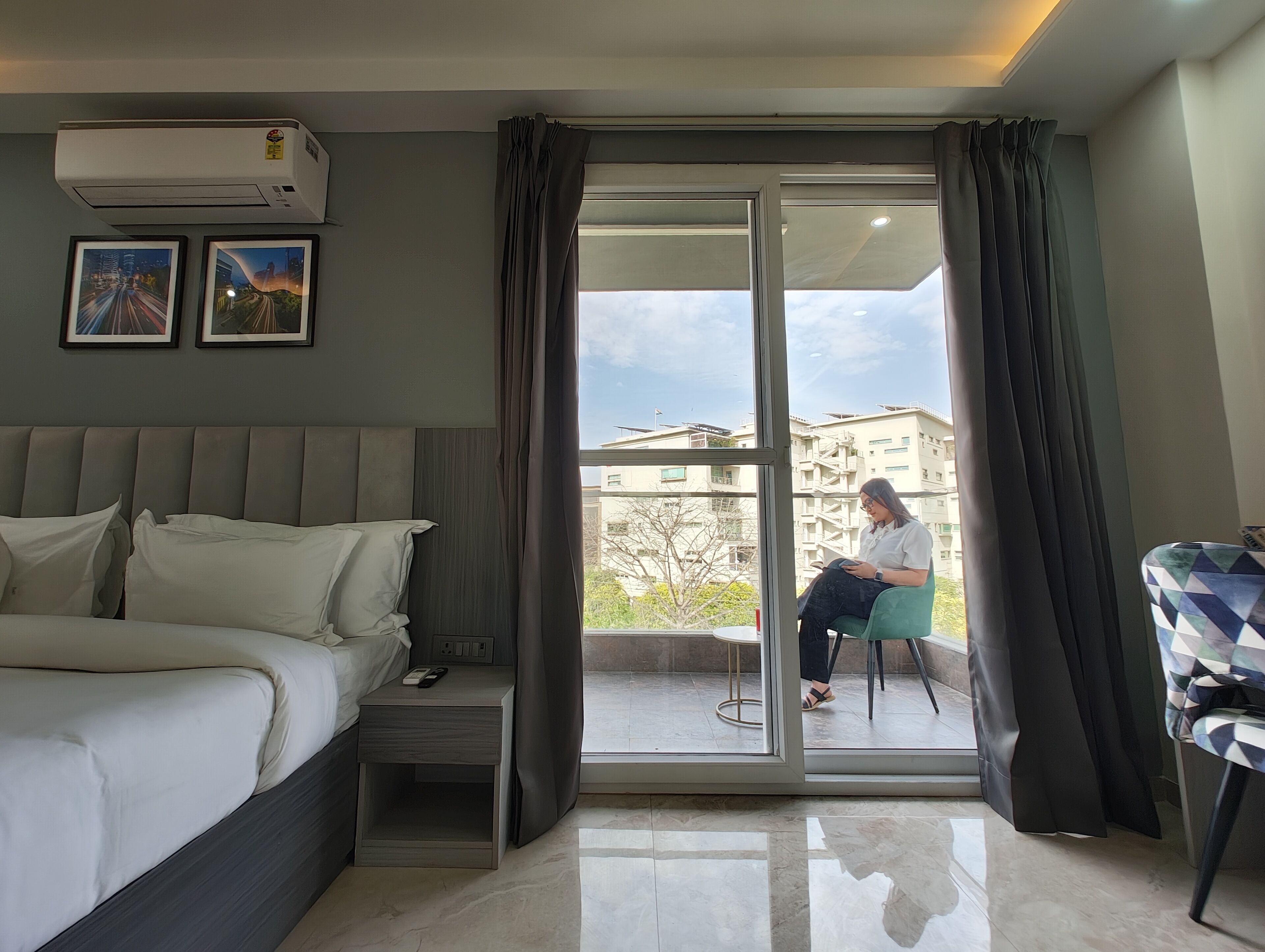 Habitación de hotel con cama, mesita de noche, una silla y un balcón con vistas a edificios.