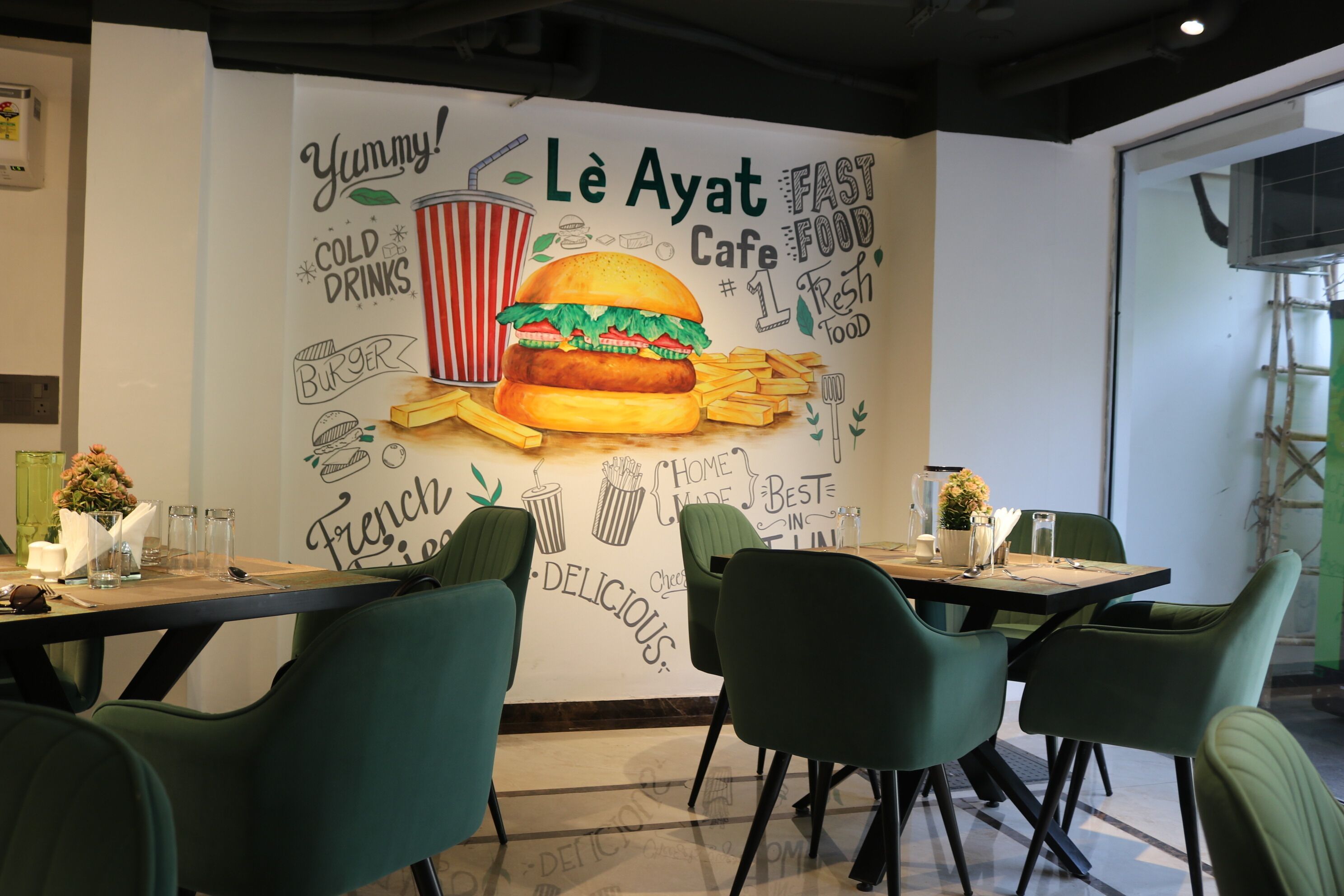 L’intérieur d’un restaurant, avec une fresque murale représentant un hamburger et des frites, des chaises vertes et des tables dressées pour le service.