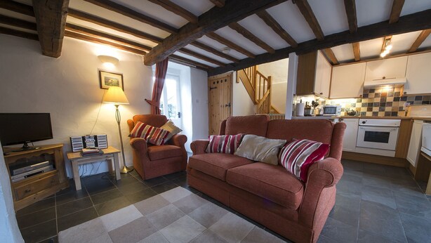 A cozy living room with a sofa, armchair, and a TV stand. A kitchen is visible in the background with a tiled backsplash and wooden cabinets. - Tarn Foot Cottage, Duddon Valley, Lake District  - A rural retreat set deep within the spectacular D (Seathwaite)