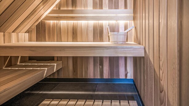 A sauna with wooden walls and benches, a bench with a bucket, and a black floor.