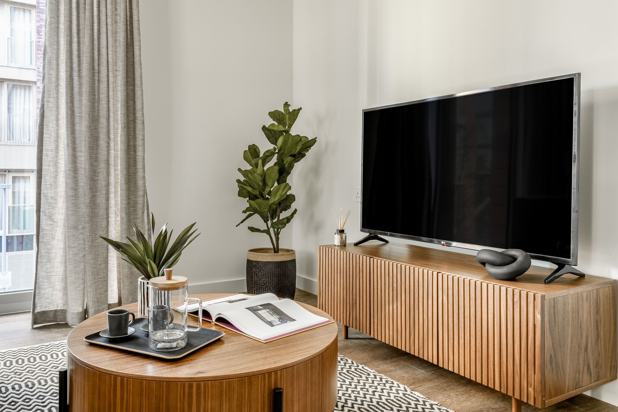 Ein modernes Wohnzimmer mit einem runden Holztisch, einem Flachbildfernseher auf einem Holzständer, einer Topfpflanze und einem großen Fenster mit Vorhängen.