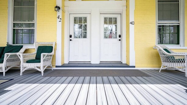 Eine Veranda mit zwei weißen Türen, einem gelben Anstrich und Korbstühlen mit grünen Kissen.