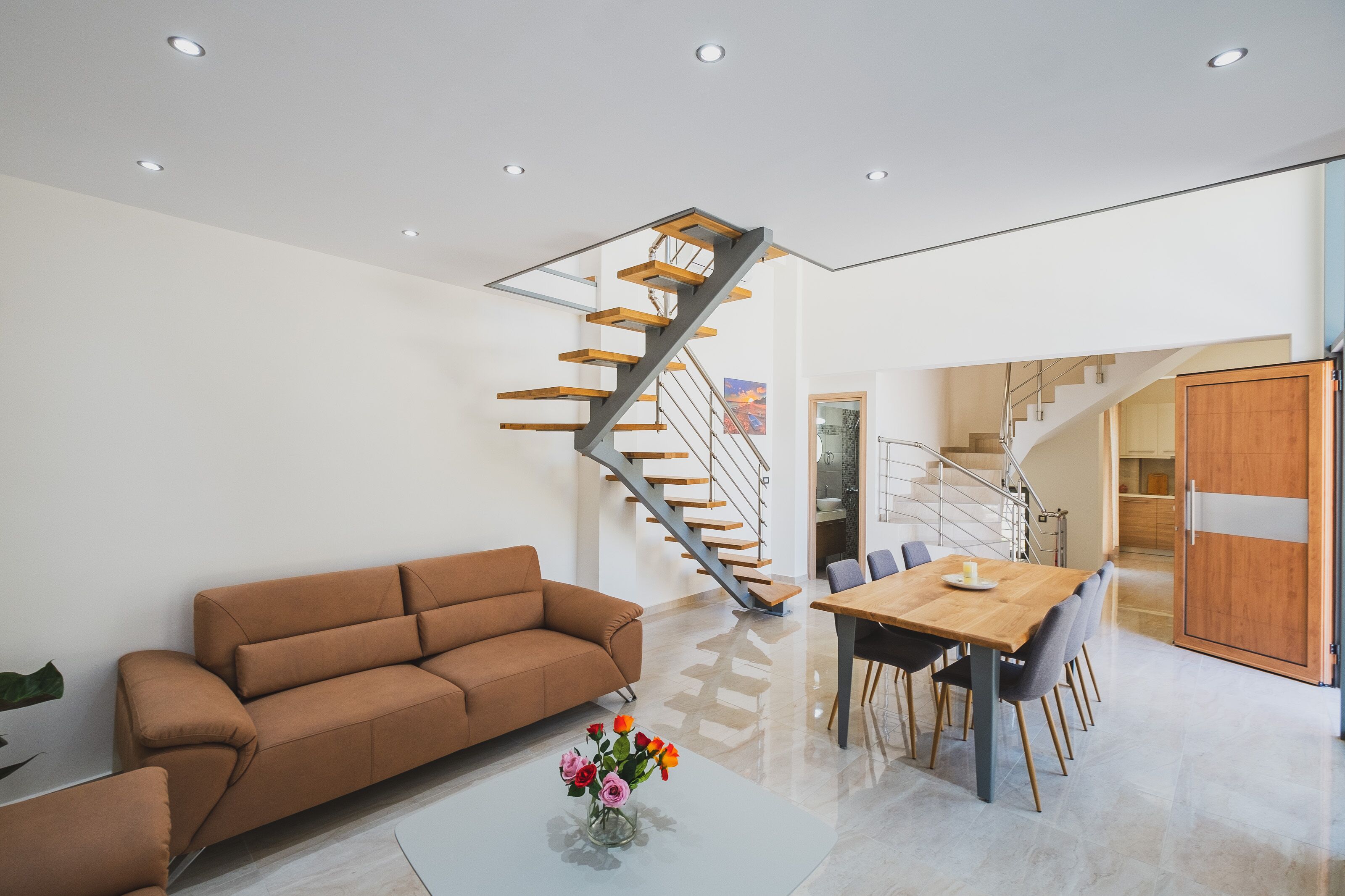 A modern living room with a brown sofa, a wooden dining table, and a staircase with wooden steps.