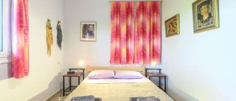 Une chambre à coucher comprenant un lit, deux tables de chevet, une commode, une fenêtre avec des rideaux et des œuvres d’art murales.