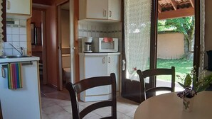 Cocina pequeña con gabinetes blancos, microondas y cafetera. Cuenta con un comedor con dos sillas y una mesa, y un ventanal corredero que da a un patio con vistas a zonas verdes.