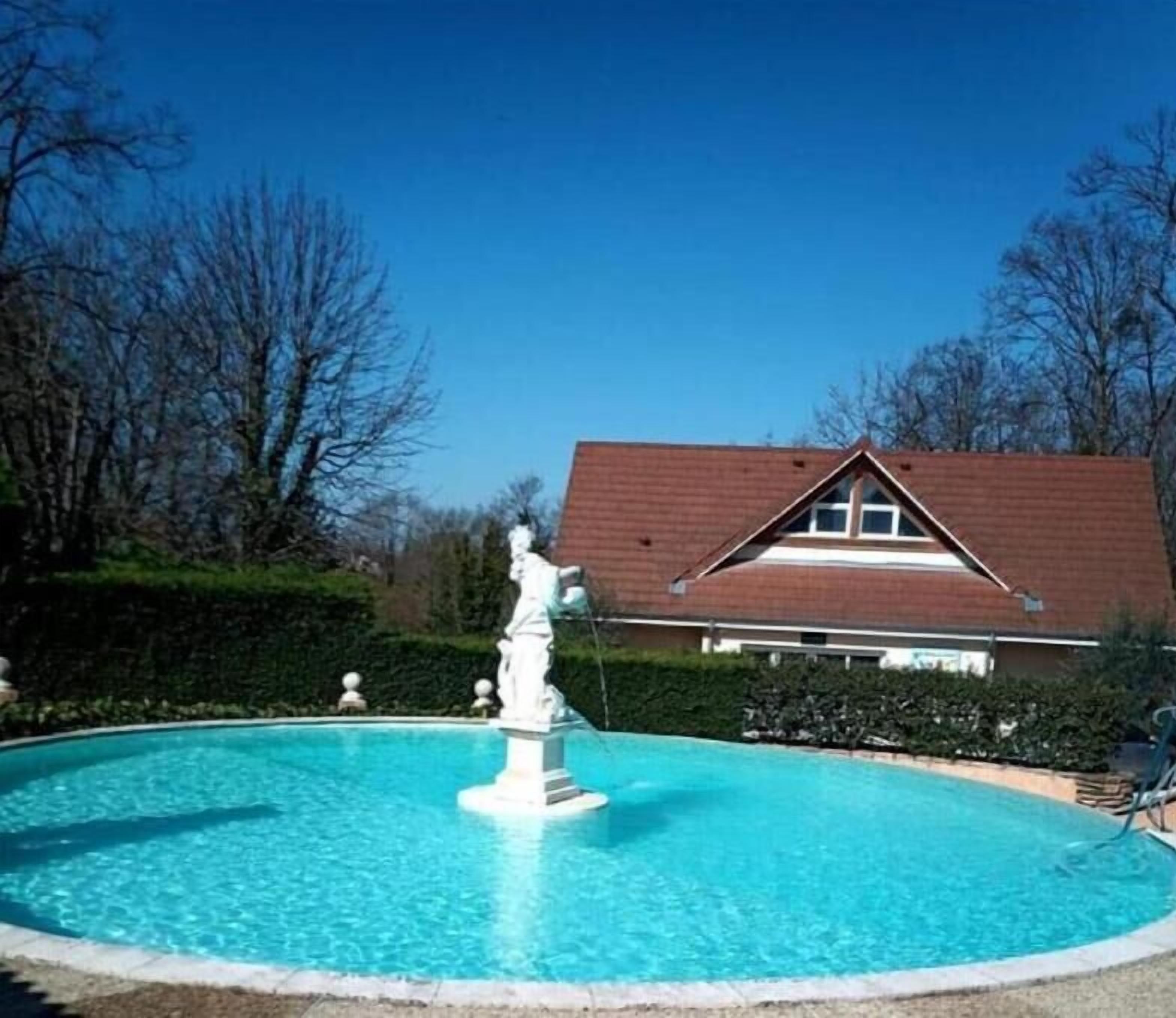 A swimming pool with a statue in the center, surrounded by a hedge, with a house in the background.