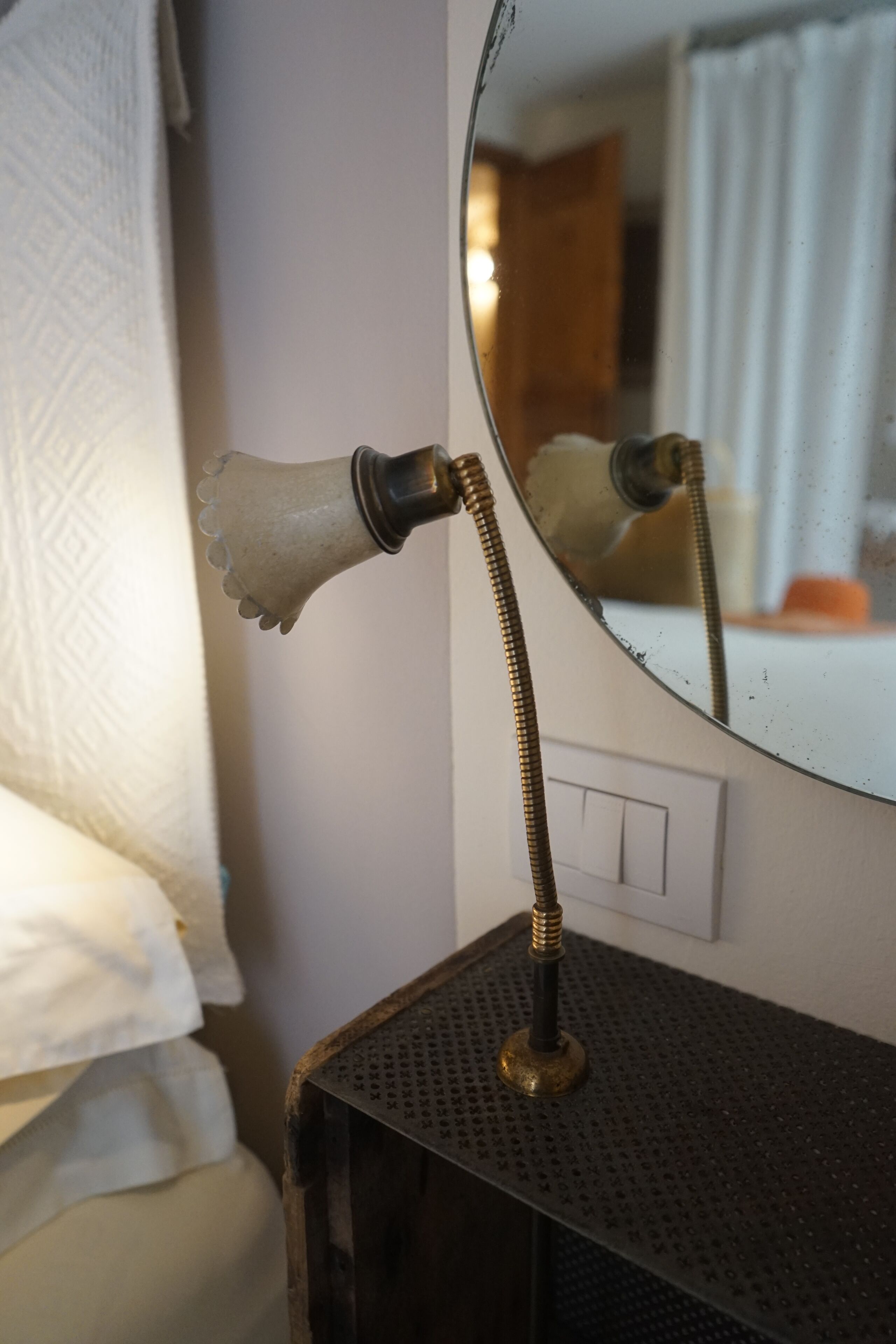 A round mirror with a brass frame and a white lampshade, reflecting a bathroom with a shower curtain and a towel.