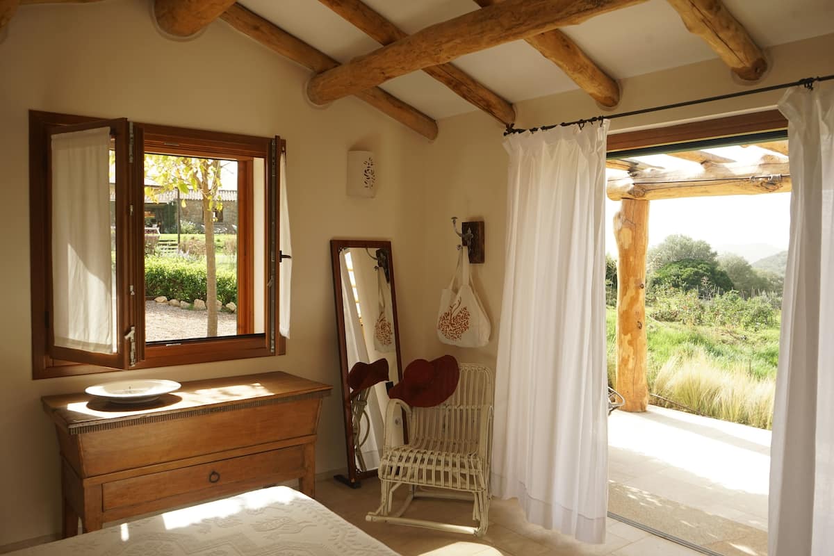 Een slaapkamer met een houten dressoir, een spiegel, een stoel en uitzicht op een tuin door een raam.