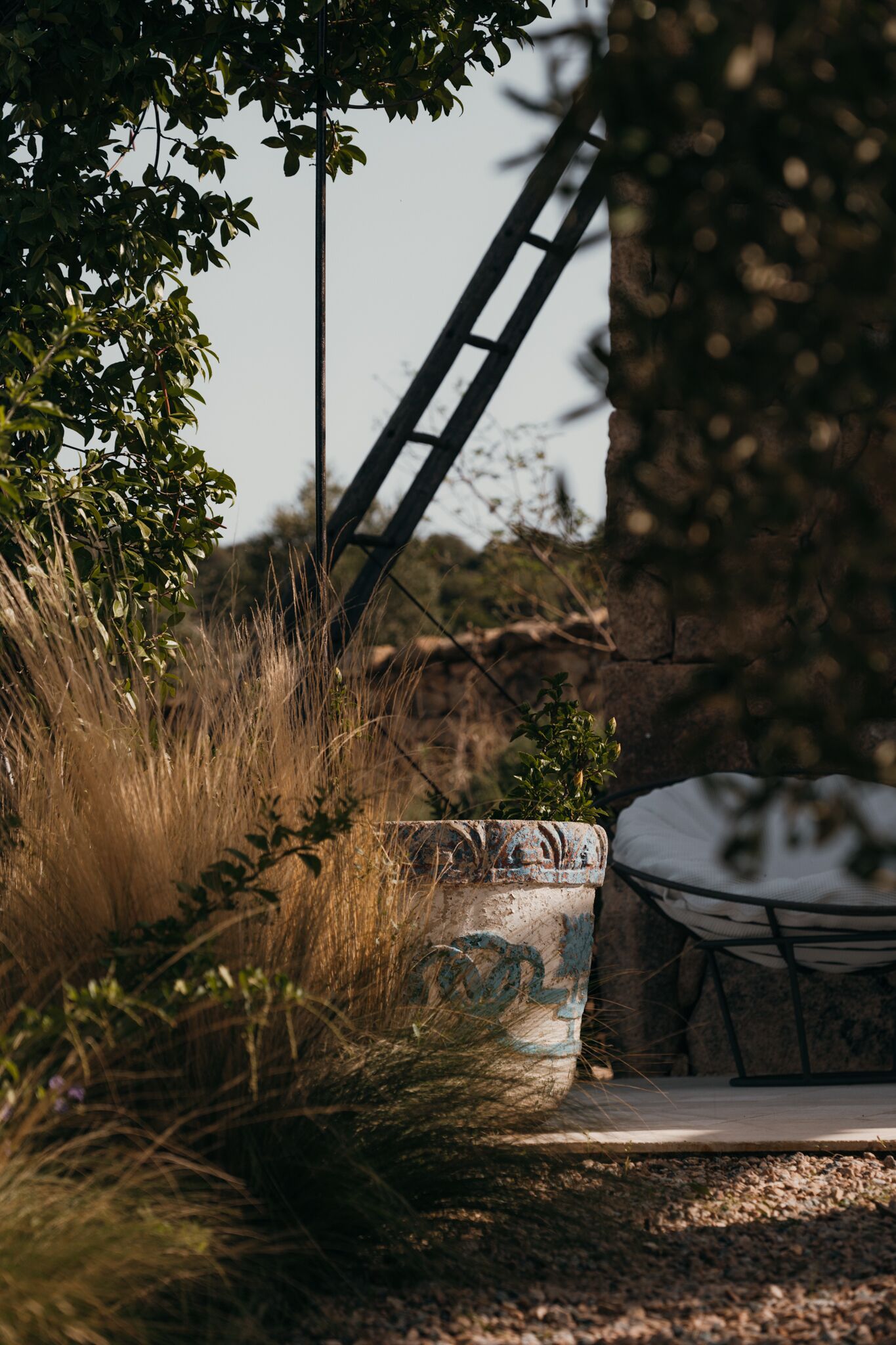 Un espace extérieur aménagé dans un style rustique, avec une chaise en métal, un bac à fleurs patiné contenant une plante, et un mur de pierre en arrière-plan.