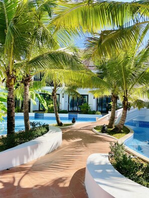 A pathway leading to a pool area surrounded by palm trees and a building with a tiled blue and white exterior. - Manyo Hotel and Resort (Luang Prabang)