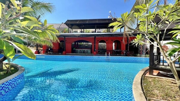 A swimming pool with a tiled edge, surrounded by palm trees and a building with red arches in the background. - Manyo Hotel and Resort (Luang Prabang)