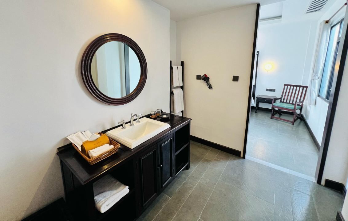 A bathroom with a large mirror, a sink, and a towel rack. There is a bench in the adjacent room.
