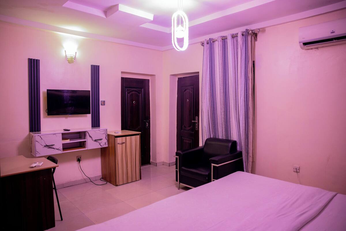 A hotel room with a bed, a desk with a chair, a TV mounted on the wall, a wardrobe, and an air conditioning unit.